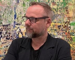 Mann mit Brille und Bart, der in einem schwarzen Blazer gekleidet ist, steht vor einer bunten, abstrakten Wandmalerei.