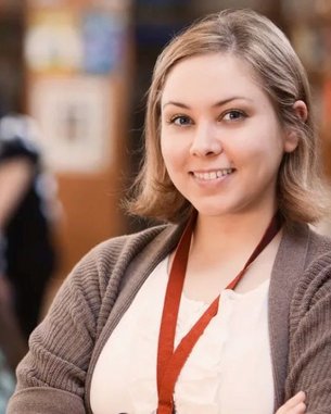 A young woman with short blonde hair is smiling in a library. In the background, other people can be seen working on computers and browsing through books.