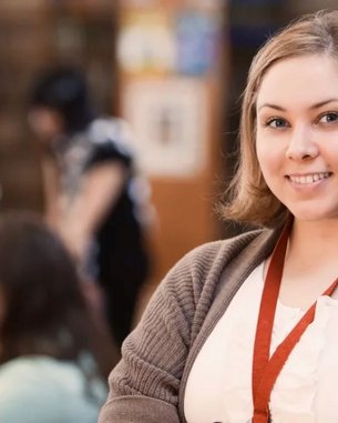 A young woman with short blonde hair is smiling in a library. In the background, other people can be seen working on computers and browsing through books.