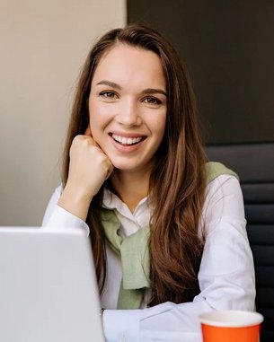 A smiling woman sits at a desk with a laptop and a cup of coffee. In the background, there’s a screen displaying text. She is wearing a white shirt and a green sweater.