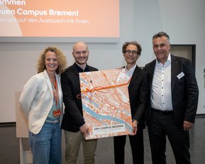 Vier Personen stehen in einem modernen Raum und halten eine große Karte von Bremen. Die Gruppe besteht aus zwei Männern und zwei Frauen, die lächeln und in formeller Kleidung gekleidet sind. Im Hintergrund ist ein Willkommensbanner sichtbar.