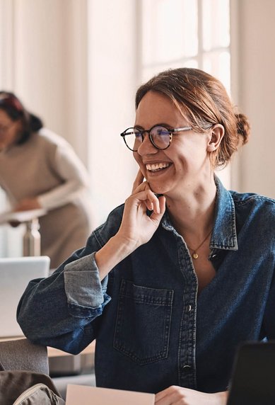 A woman with glasses sits at a table, smiling as she thoughtfully plays with her hand on her face. In the background, several people are busy with their laptops.