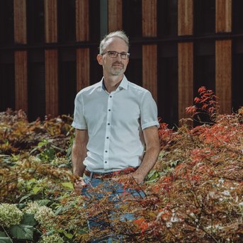 Ein Mann steht in einem Garten mit buntem Laub und Pflanzen. Er trägt ein weißes Hemd und eine graue Hose. Im Hintergrund sind Holzpaneele eines Gebäudes sichtbar.