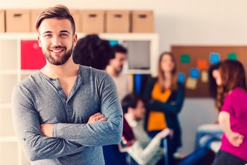 Ein Mann mit Bart lächelt und steht mit verschränkten Armen im Vordergrund, während eine Gruppe von Menschen im Hintergrund miteinander spricht.