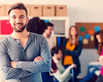 Ein Mann mit Bart lächelt und steht mit verschränkten Armen im Vordergrund, während eine Gruppe von Menschen im Hintergrund miteinander spricht.