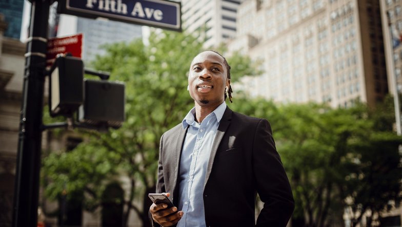 Kategorie New Work Studium studieren SRH University Ein Mann in einem Anzug steht an einer Straßenecke mit einem Smartphone in der Hand. Im Hintergrund ist ein Straßenschild mit der Aufschrift "Fifth Ave" und grüne Bäume zu sehen.