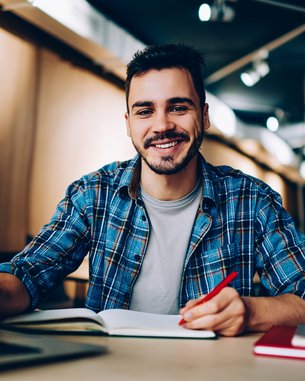 A young man with a beard is sitting at a table, smiling and looking at the camera. He is wearing a checked shirt and holding a red pen. In front of him, there is a notebook and a laptop.