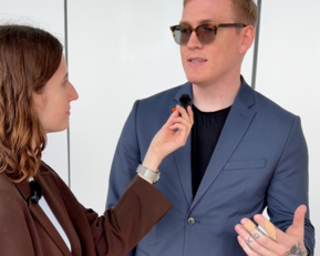 Eine Frau interviewt einen Mann in einem Anzug, während sie ein Mikrofon an seinem Kragen befestigt. Beide schauen sich an.