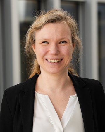 A smiling woman with blonde, curly hair is wearing a black jacket over a white top.