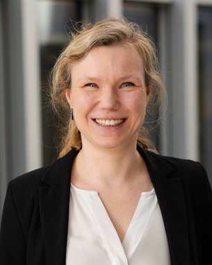 A smiling woman with blonde, curly hair is wearing a black jacket over a white top.