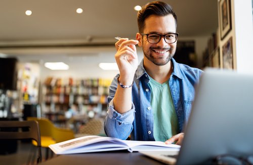 A man with glasses sits at a table, smiling and holding a pen while looking at a laptop and taking notes.