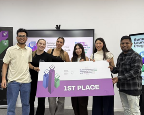 Sechs Personen stehen zusammen und halten ein großes Schild mit der Aufschrift "1ST PLACE" während einer Preisverleihung im Rahmen eines Programms.