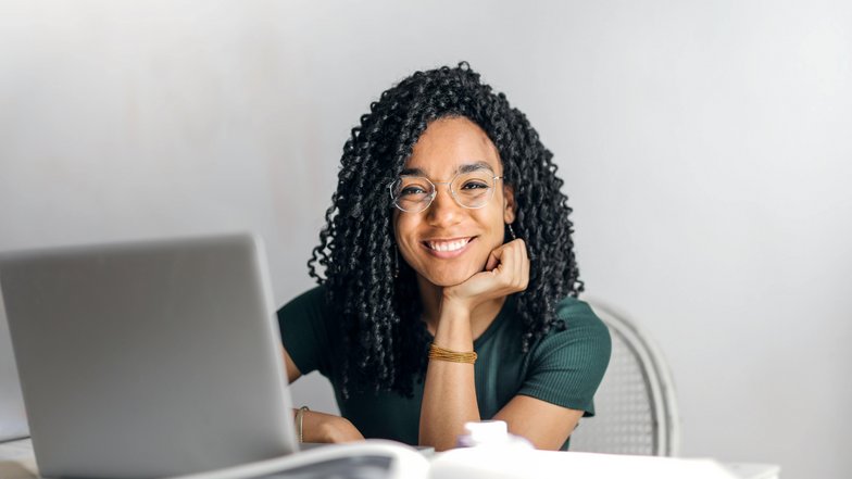 Kategorie Informatik SRH University A smiling woman with curly hair sits at a table in front of a laptop, looking kindly at the camera. She is wearing a green blouse and glasses.