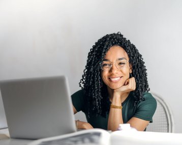 Kategorie Informatik SRH University A smiling woman with curly hair sits at a table in front of a laptop, looking kindly at the camera. She is wearing a green blouse and glasses.
