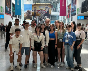 A group of people is posing in a large hall with colorful banners and people in the background.