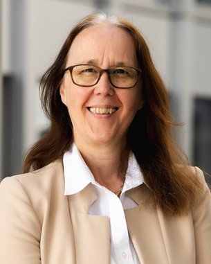 A woman with long brown hair and glasses smiles warmly. She is wearing a light blazer and a white blouse.