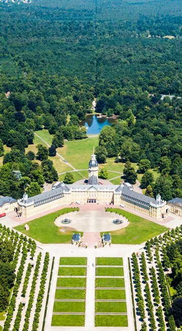 Luftaufnahme eines großen Parks mit symmetrischen Gartenanlagen, umgeben von Bäumen und Gebäuden. Im Hintergrund erstreckt sich eine grüne Landschaft bis zum Horizont.