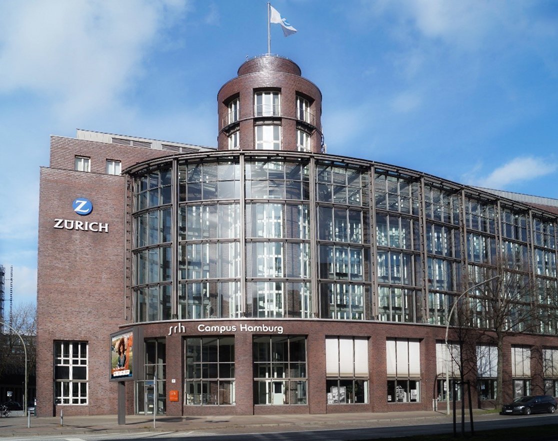 Modern glass facade of the SRH Campus Homburg featuring the Zurich Insurance logo. The building has a round tower and is surrounded by a bright environment.