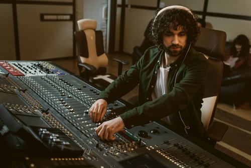 A man with curly hair sits at a mixing console, adjusting the sliders in a recording studio. In the background, other people can be seen.
