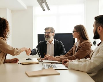 Kategorie Management SRH University A business meeting where a woman shakes hands with a man. In the background, two other people are seated at a table with laptops and notes. A bright office environment with natural light.