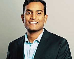 A man with dark hair is wearing a black suit and a light blue shirt. He is smiling and standing in front of a neutral background. His pose is friendly and professional.