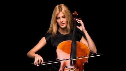 A young woman is playing the cello while intently looking at the strings. She has long blonde hair and is wearing a black top.