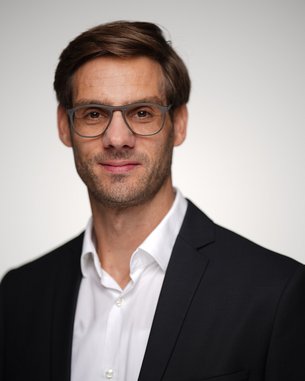 Ein Mann mit Brille und Anzug lächelt in die Kamera. Der Hintergrund ist unscharf und hell, was den Fokus auf sein Gesicht lenkt.