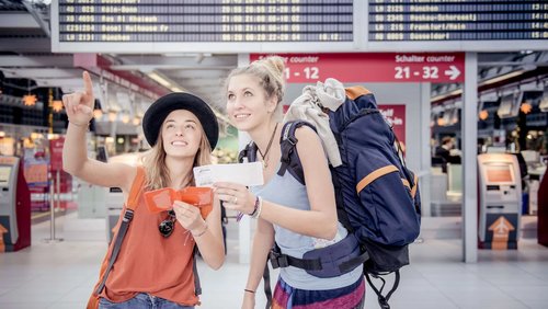 Zwei junge Frauen stehen in einem Flughafen und schauen auf eine Anzeigetafel. Eine Frau zeigt mit dem Finger nach oben, während die andere ein Ticket in der Hand hält. Beide tragen Rucksäcke und lächeln.