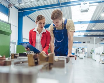 A woman in a red blazer and a man in blue work clothes are examining parts together on a table in a workshop.