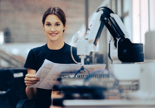 A woman is holding documents and smiling, while a robotic arm stands next to her. She is in a modern workplace.