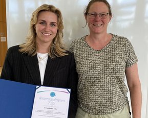Zwei Frauen stehen nebeneinander, eine hält ein Zertifikat für den Wissenschaftspreis 2025 in der Hand. Beide lächeln in die Kamera.
