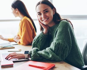 Eine junge Frau mit langen Haaren sitzt an einem Tisch, lächelt und arbeitet an einem Laptop. Im Hintergrund sind weitere Personen und Laptops zu sehen.