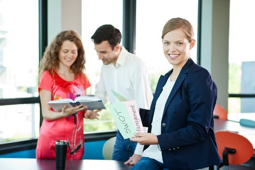 Drei Personen in einem modernen Büro, eine Frau hält ein Blatt mit der Aufschrift "SWOT Management", während die anderen beiden ein Buch betrachten.