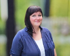 Eine Frau mit dunklen Haaren trägt ein blaues Hemd und steht lächelnd vor einem unscharfen, grünen Hintergrund.