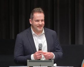 Ein Mann in einem Anzug hält ein Mikrofon und spricht auf einer Bühne. Im Hintergrund sind Vorhänge und ein orangefarbenes Logo sichtbar.