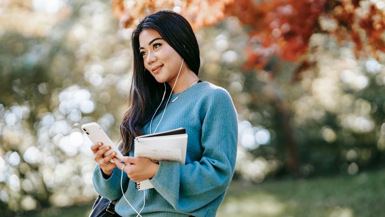 Eine junge Frau mit langen, dunklen Haaren steht im Freien. Sie trägt einen blauen Pullover, hört Musik über Kopfhörer und hält ein Smartphone sowie Notizblätter in der Hand. Bunte Herbstblätter sind im Hintergrund sichtbar.