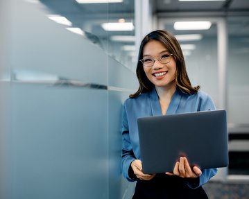 A woman with glasses is holding a laptop in a modern office and smiling warmly. She is wearing a blue blouse.