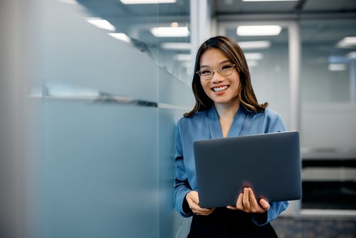 A woman with glasses is holding a laptop in a modern office and smiling warmly. She is wearing a blue blouse.