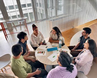 Six people are sitting around a table discussing. On the table, there are documents and drinks. The room has large windows and a modern design.