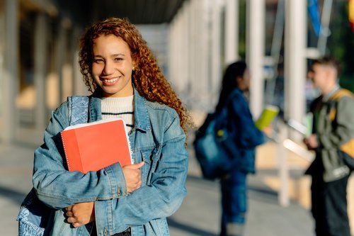 Eine junge Frau mit lockigem, braunem Haar trägt eine Jeansjacke und hält ein rotes Notizbuch. Im Hintergrund sind weitere Studierende zu sehen.