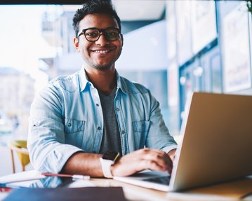 A young man with glasses is sitting at a table in a café, working on a laptop. He is smiling warmly.