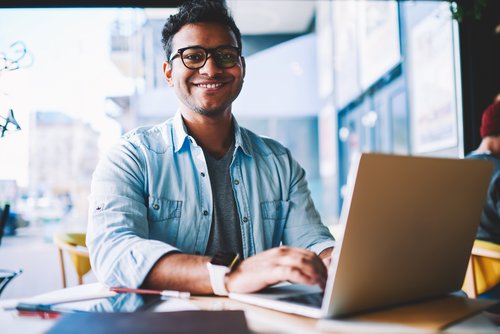 A young man with glasses is sitting at a table in a café, working on a laptop. He is smiling warmly.