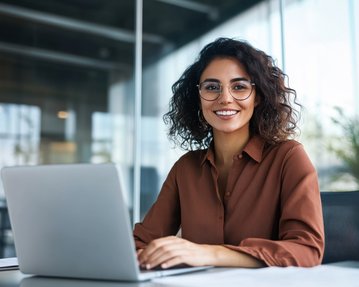 A woman with curly hair and glasses sits at a desk, smiling as she works on a laptop.