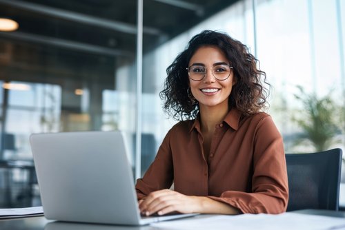 A woman with curly hair and glasses sits at a desk, smiling as she works on a laptop.
