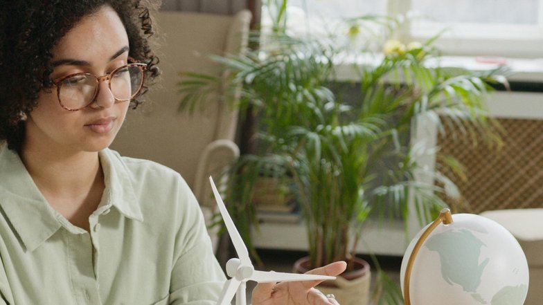 Kategorie Nachhaltigkeit studieren Studium SRH University A woman with curly hair and glasses sits at a table, pointing at a model of a wind turbine and small houses, while a globe stands in the background. Plants are also visible.