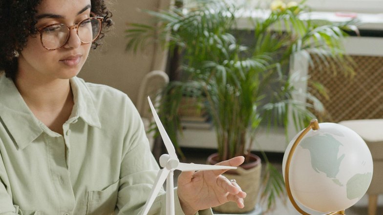 Kategorie Nachhaltigkeit studieren Studium SRH University A woman with curly hair and glasses sits at a table, pointing at a model of a wind turbine and small houses, while a globe stands in the background. Plants are also visible.