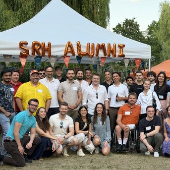 Group meeting of SRH alumni under a tent, with participants in various outfits, smiling and standing around a banner decoration.