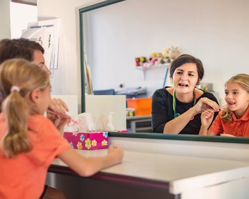 Eine Frau erklärt einem Mädchen im orangefarbenen Shirt etwas vor einem Spiegel. Das Mädchen hält eine Zahnbürste in der Hand. Auf dem Tisch steht eine Box mit Taschentüchern und Spielzeug im Hintergrund.