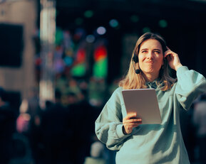 A young woman with headphones is holding a tablet in her hand and smiling as she stands in front of a stage with people.