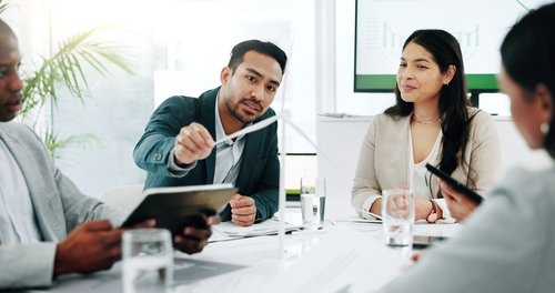 A man is presenting during a meeting while three other people listen attentively. Graphics are visible in the background.
